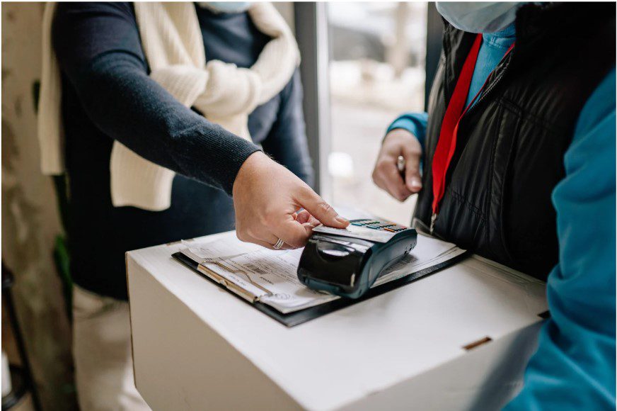 A customer taps a credit card on a portable payment terminal held by a delivery worker during a package drop-off.