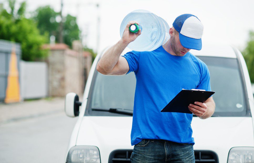 Water delivery driver carrying a large bottle and checking orders on a clipboard in front of a delivery van