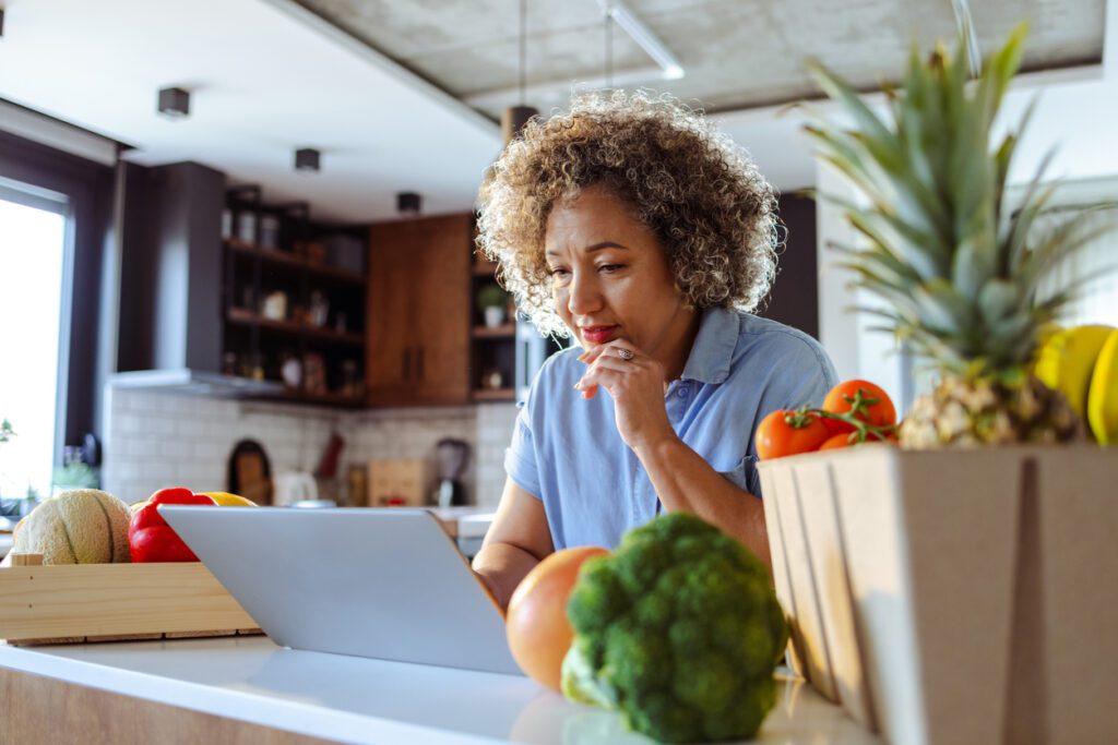 Woman confirms her next produce order from her laptop surrounded by items from her current order. 