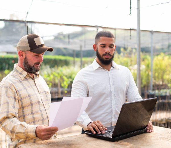 Two farmers stand in a greenhouse looking at farming accounting software on their laptop.