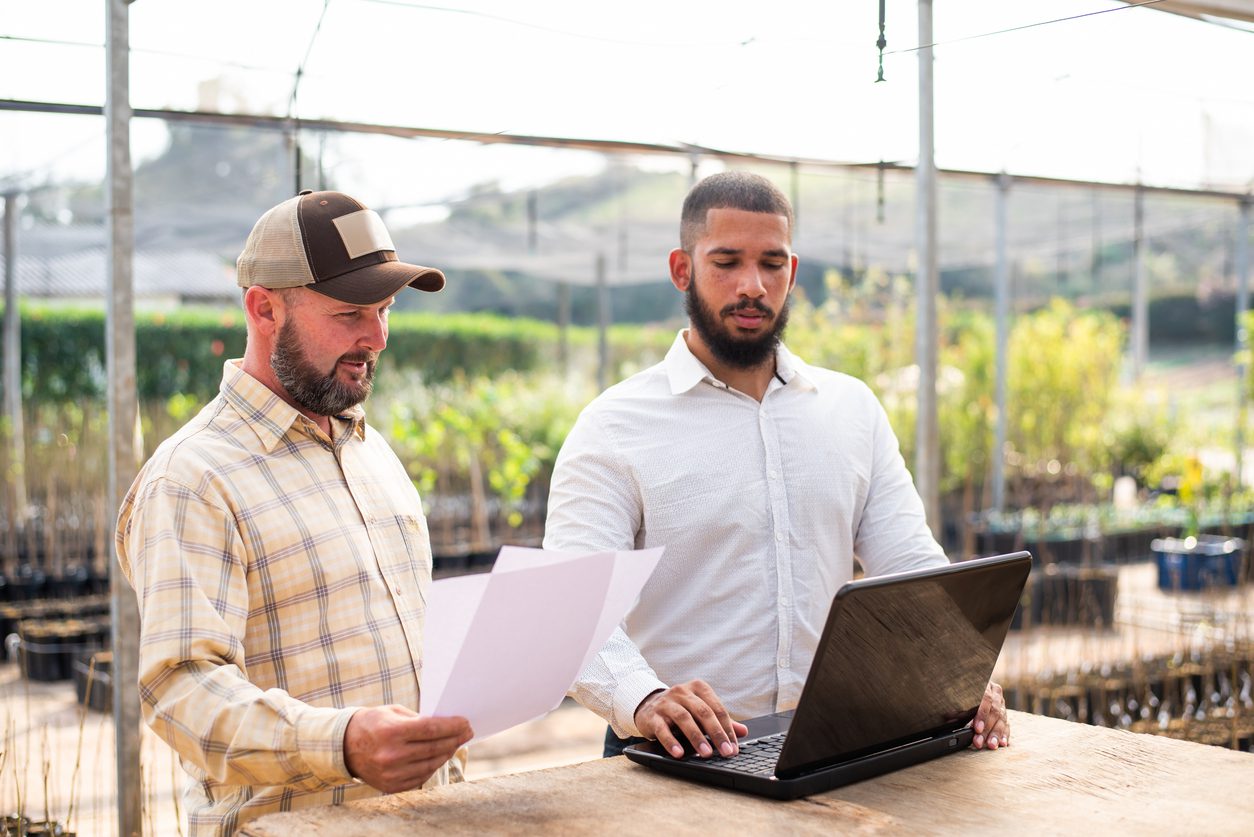 Two farmers stand in a greenhouse looking at farming accounting software on their laptop.